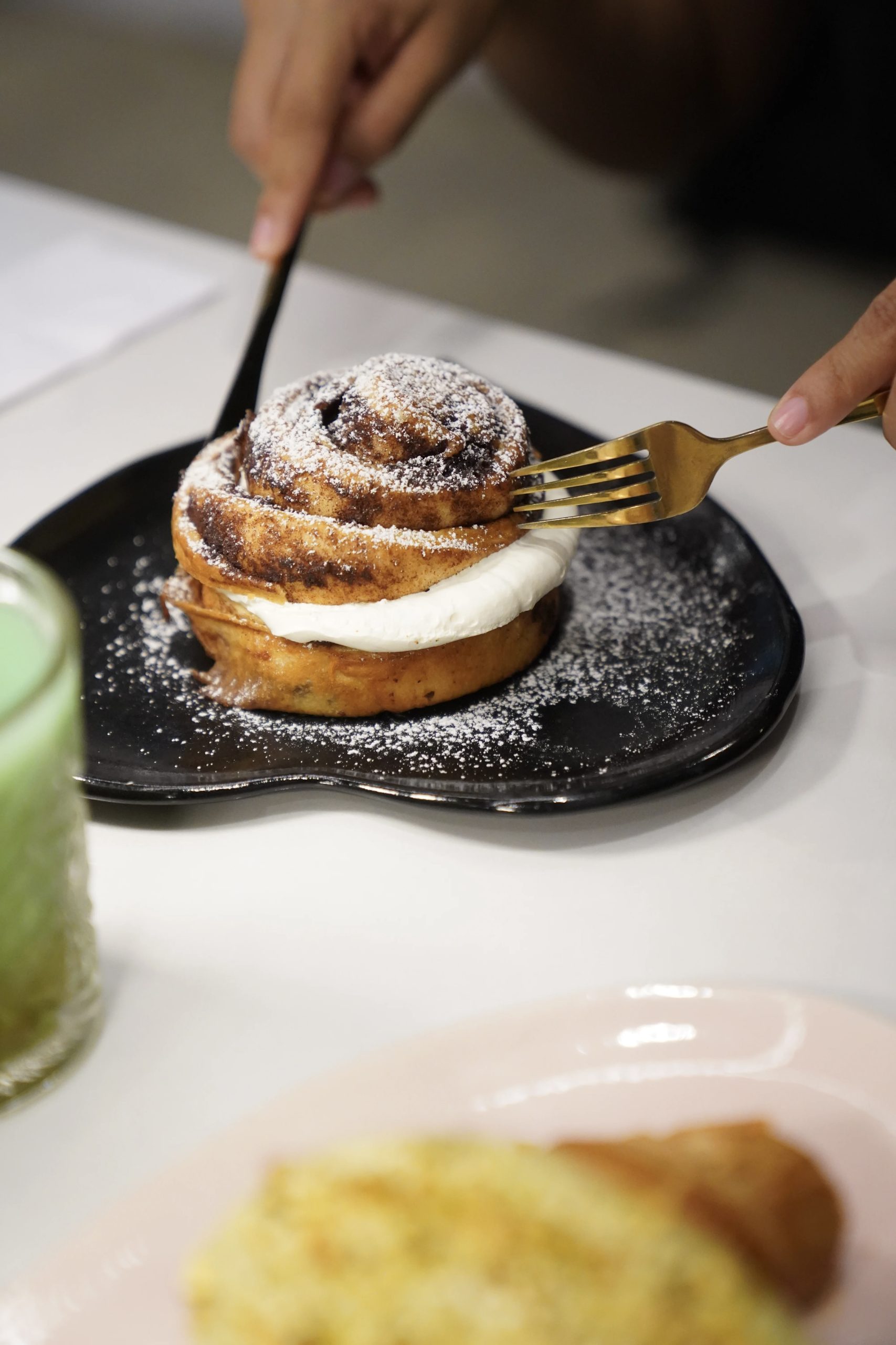 Cinnamon roll con azúcar pulverizada servido en plato negro, cortado con tenedor y cuchillo dorados – fotografía editorial de ambiente de café por Suma Estudios