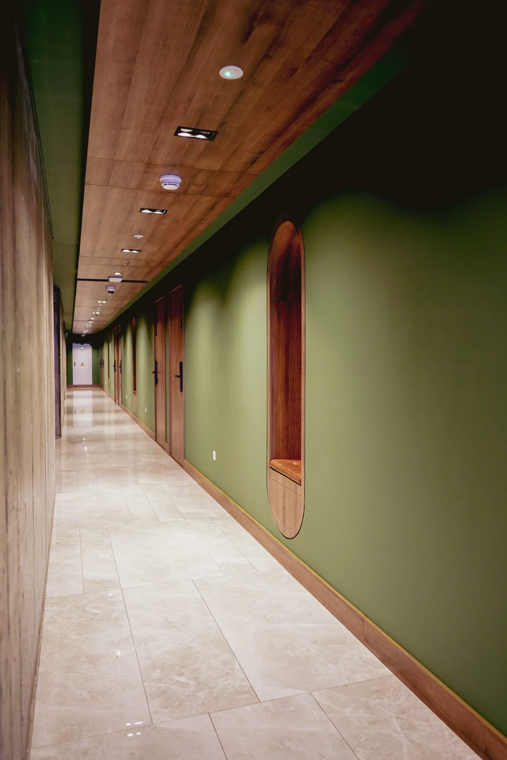 Pasillo largo con paredes verde oliva, techo de madera oscura y piso de mármol claro – diseño interior con nicho de asiento empotrado – fotografía por Suma Estudios