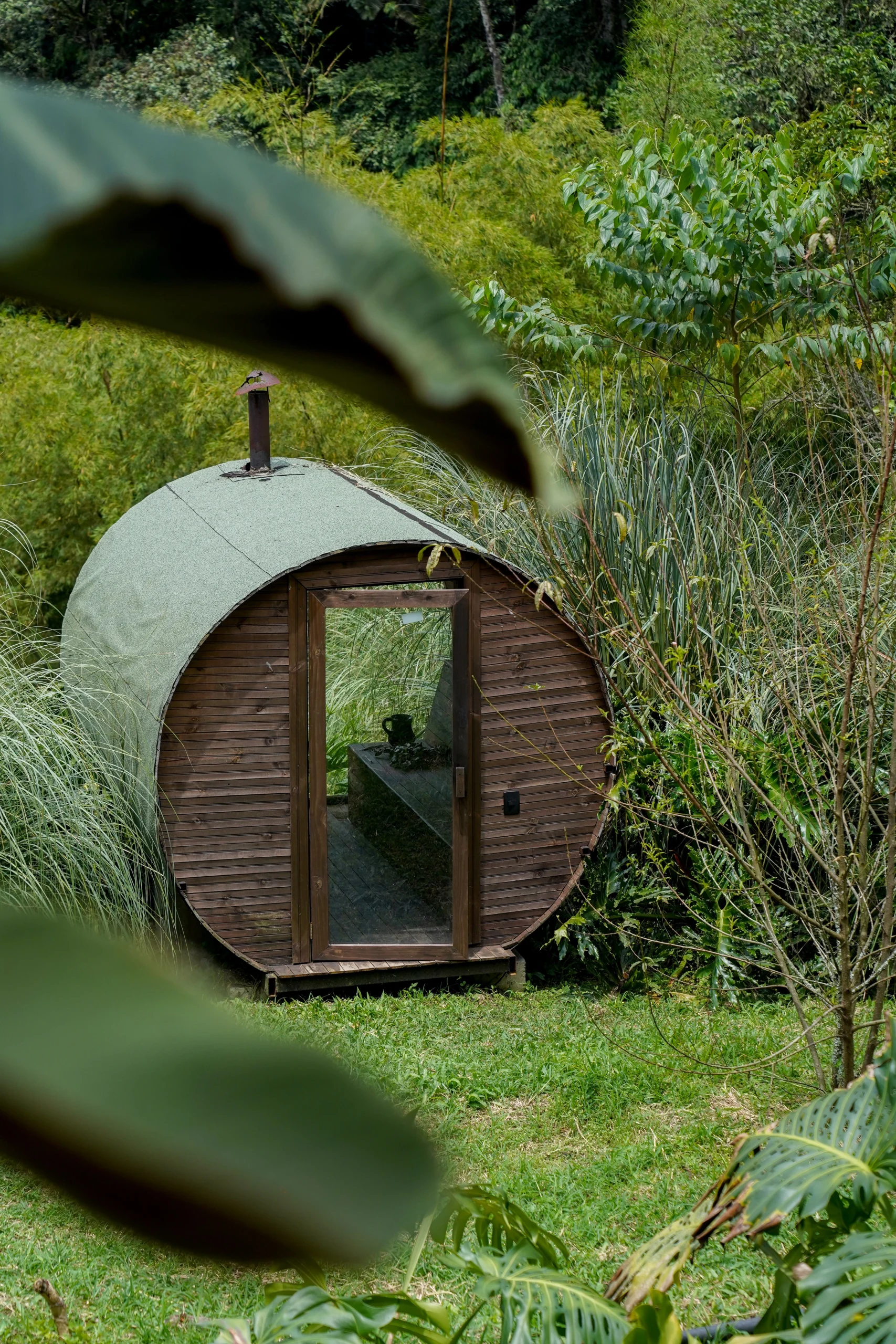 Sauna cilíndrica de madera en entorno verde – estructura rústica con techo abovedado y puerta de vidrio – fotografía de arquitectura integrada por Suma Estudios