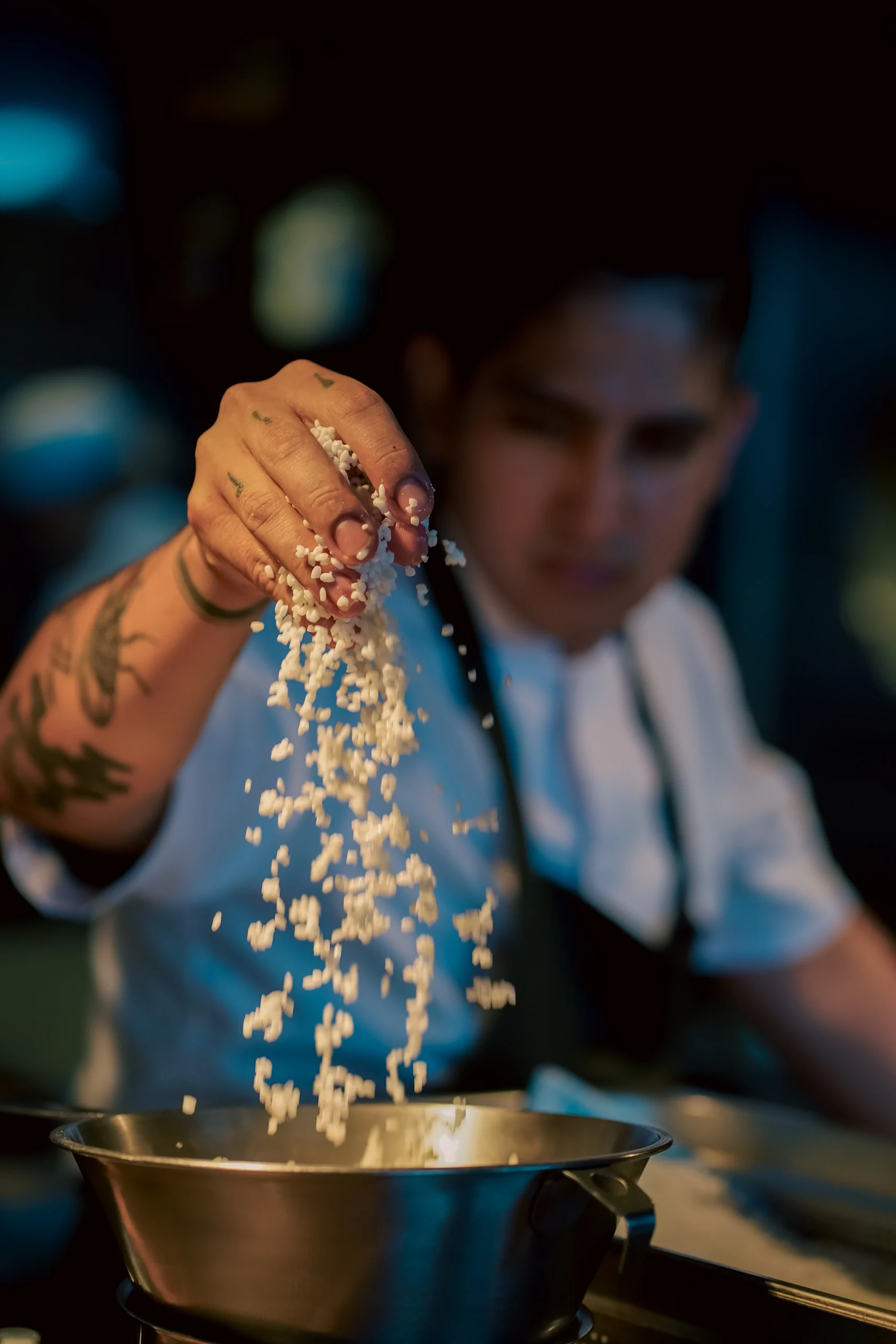 Chef con tatuajes añadiendo arroz blanco a sartén metálica – primer plano con enfoque en ingredientes – fotografía gastronómica por Suma Estudios