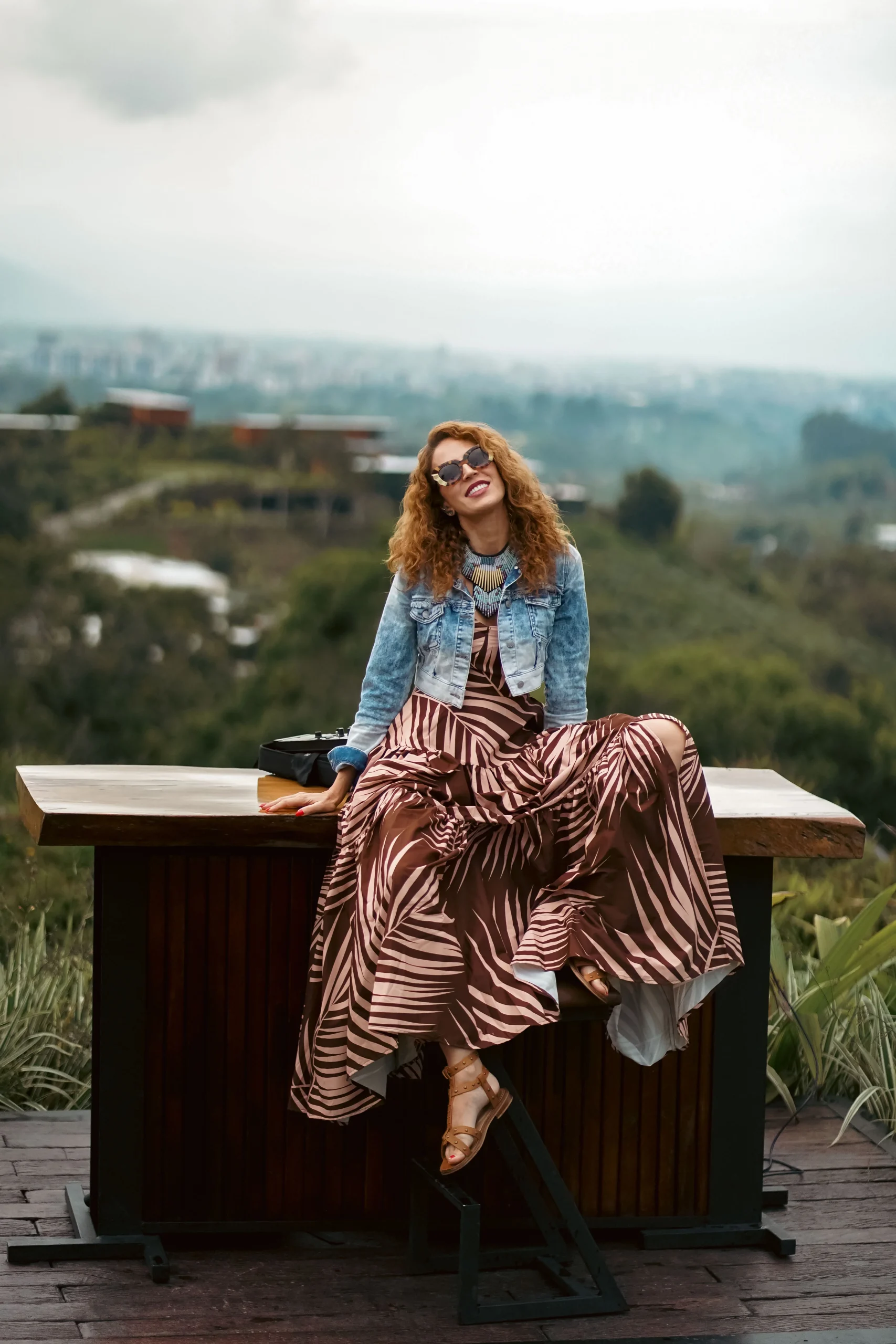 Alejandra Azcárate sentada en barra de madera al aire libre, con vestido estampado de cebra y blusa de mezclilla, contemplando una vista panorámica de colinas – fotografía editorial por Suma Estudios