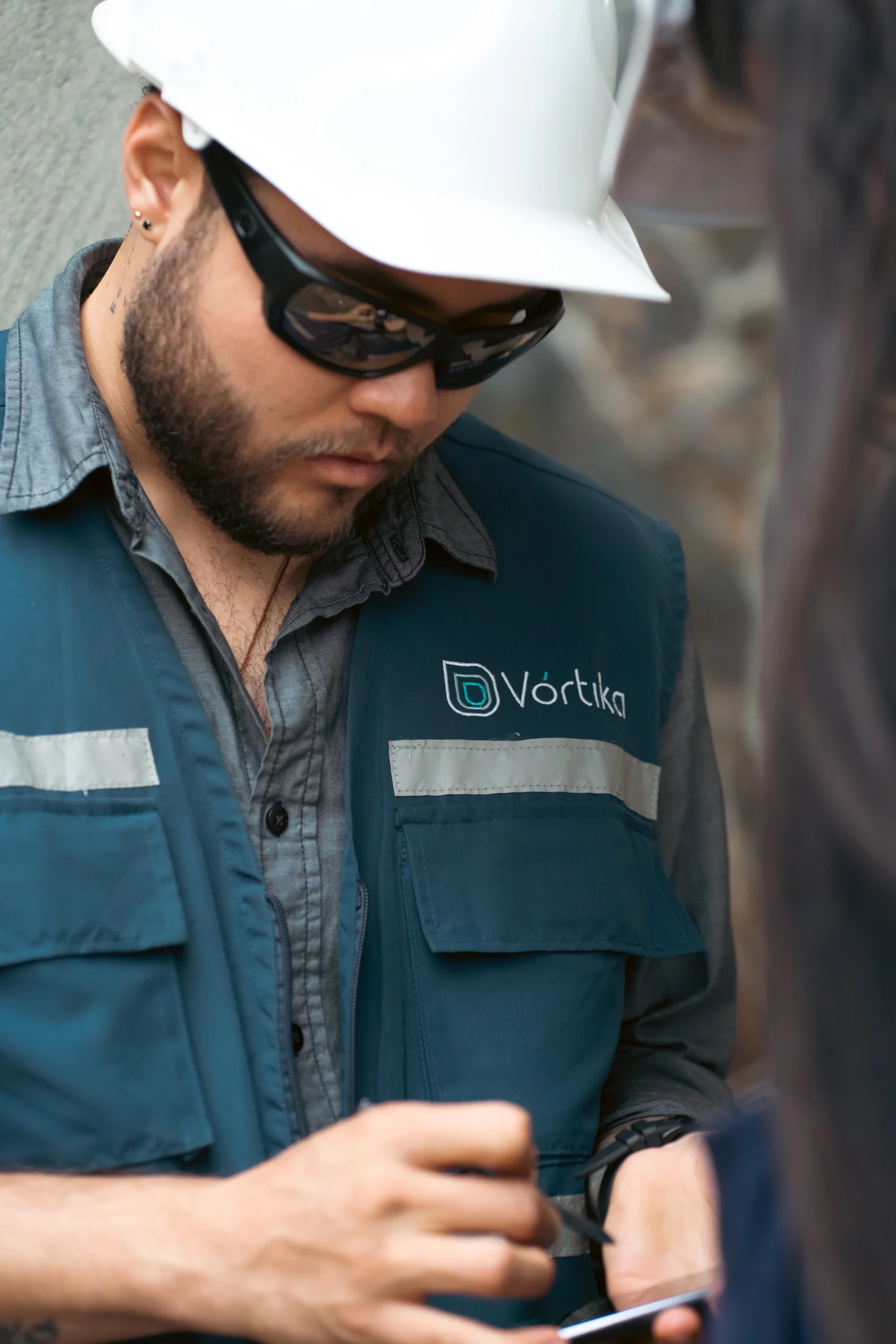 Hombre con casco blanco, gafas de protección y chaleco azul con logo de Vórtika tomando notas en sitio de trabajo – fotografía documental industrial por Suma Estudios